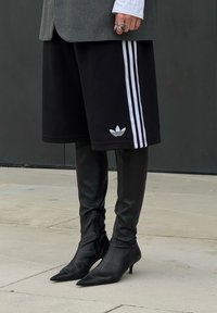 Schwarze Sweatshorts mit weißen Adidas-Streifen, kombiniert mit eng anliegenden schwarzen Leder-Stöckelschuhen. Grauer oversized Blazer und weißes Hemd sind sichtbar.
