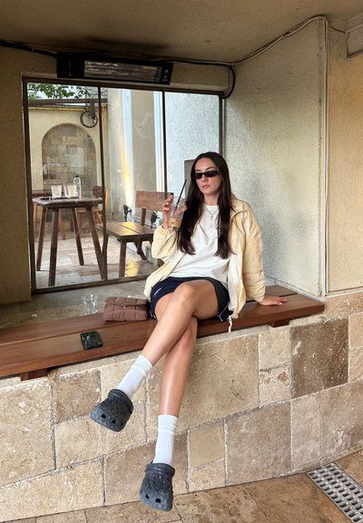 Chaqueta amarilla clara, camiseta blanca, pantalones cortos azul marino, calcetines blancos con Crocs negros. Sentado en un saliente de madera cerca de una pared de piedra, sosteniendo una bebida.