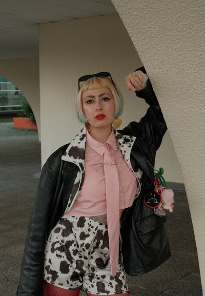 Chaqueta de cuero negra sobre una camisa rosa con una corbata, pantalones cortos de estampado de vaca y pendientes en forma de corazón. Cabello colorido, múltiples accesorios.