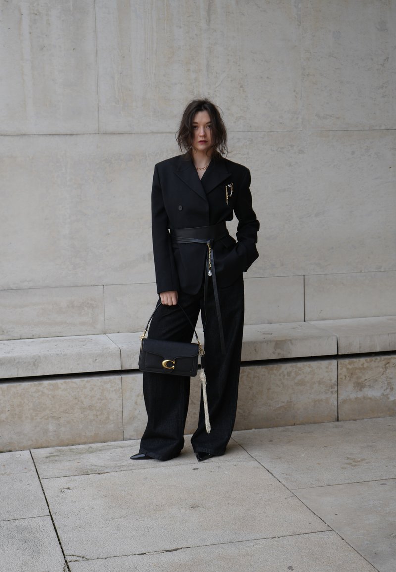 Γυναίκα που φοράει φαρδιά μαύρη σακάκα με ζώνη, παντελόνια με φαρδιά μπατζάκια, κρατάει μαύρη τσάντα και στέκεται μπροστά σε ένα φωτεινό πέτρινο τοίχο έξω.