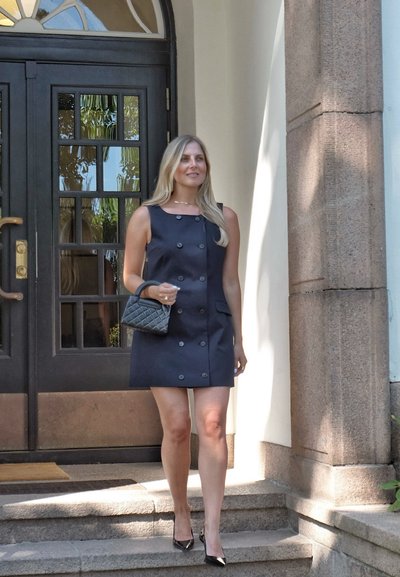 Vestido sin mangas de color azul marino con botones de doble botonadura, bolsillos planos y una silueta cuadrada; combinado con un bolso negro acolchado y tacones negros.