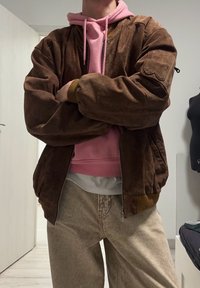 Veste bomber en daim marron avec des poignets côtelés et des accents colorés sur un hoodie rose, associée à un pantalon large beige clair.