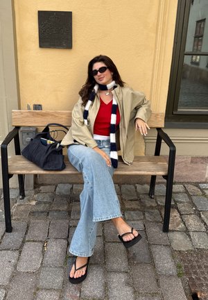 Modelo vistiendo una chaqueta beige sobre una blusa roja, pantalones vaqueros acampanados de color azul claro y chanclas negras, sentada en un banco de madera al lado de una bolsa negra.