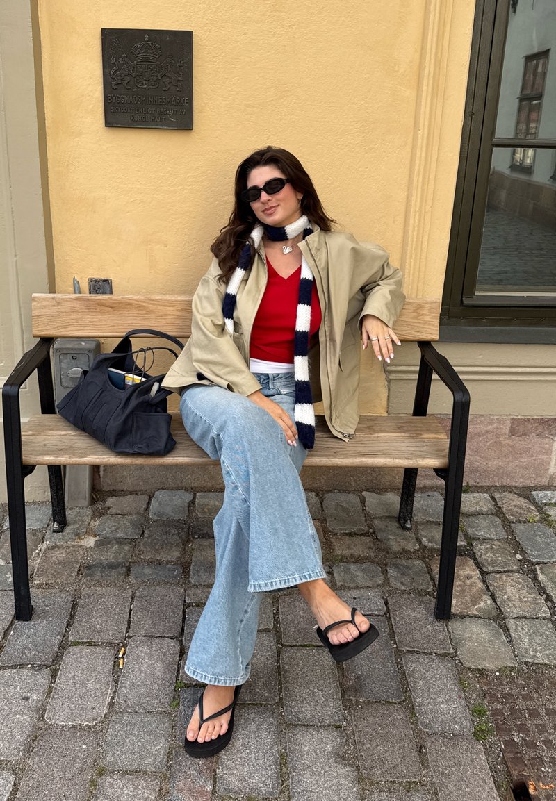 Modelo vestindo uma jaqueta bege sobre uma blusa vermelha, jeans claros flare e chinelos pretos, sentado em um banco de madeira ao lado de uma bolsa preta.