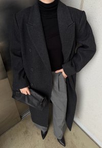Manteau croisé en laine grise avec revers à pointe, col roulé noir, pantalon plissé gris et pochette en cuir noire. Talons noirs pointus.