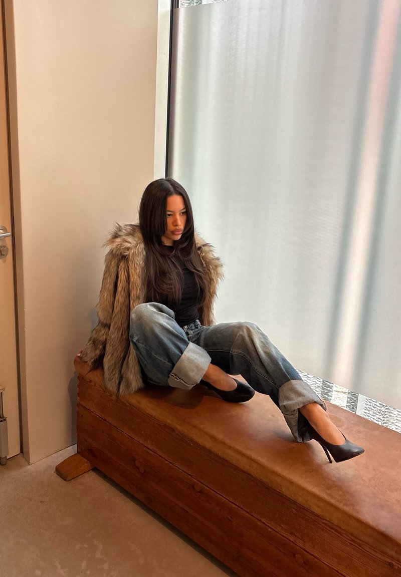 Νεαρή γυναίκα με γούνινο παλτό, μαύρο τοπ και τζιν γυρισμένο στα μπατζάκια κάθεται σε ξύλινο παγκάκι κοντά σε παράθυρο με παγωμένο γυαλί.