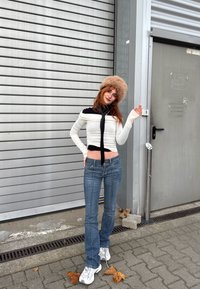 Top crop alb cu mâneci lungi, accente negre și o fundă, blugi albaștri și adidași argintii; purtând o pălărie pufoasă bej, pozând cu un gest jucăuș.