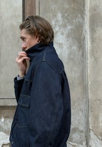 Mørk denimjakke med høj krave, sidelommer og kontrastsyninger. Glat tekstur og lige pasform.