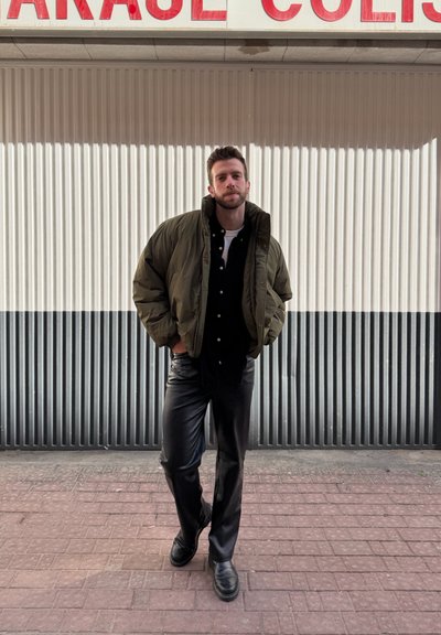 Hombre con chaqueta acolchada verde, camisa negra, pantalones de cuero y botas, de pie con las manos en los bolsillos frente a una pared corrugada en blanco y negro.