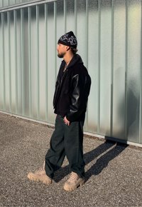 Veste en velours côtelé noire avec manches en cuir, sweat à capuche noir en dessous, pantalon vert olive et baskets à plateforme beiges. Bonnet noir avec motif blanc.