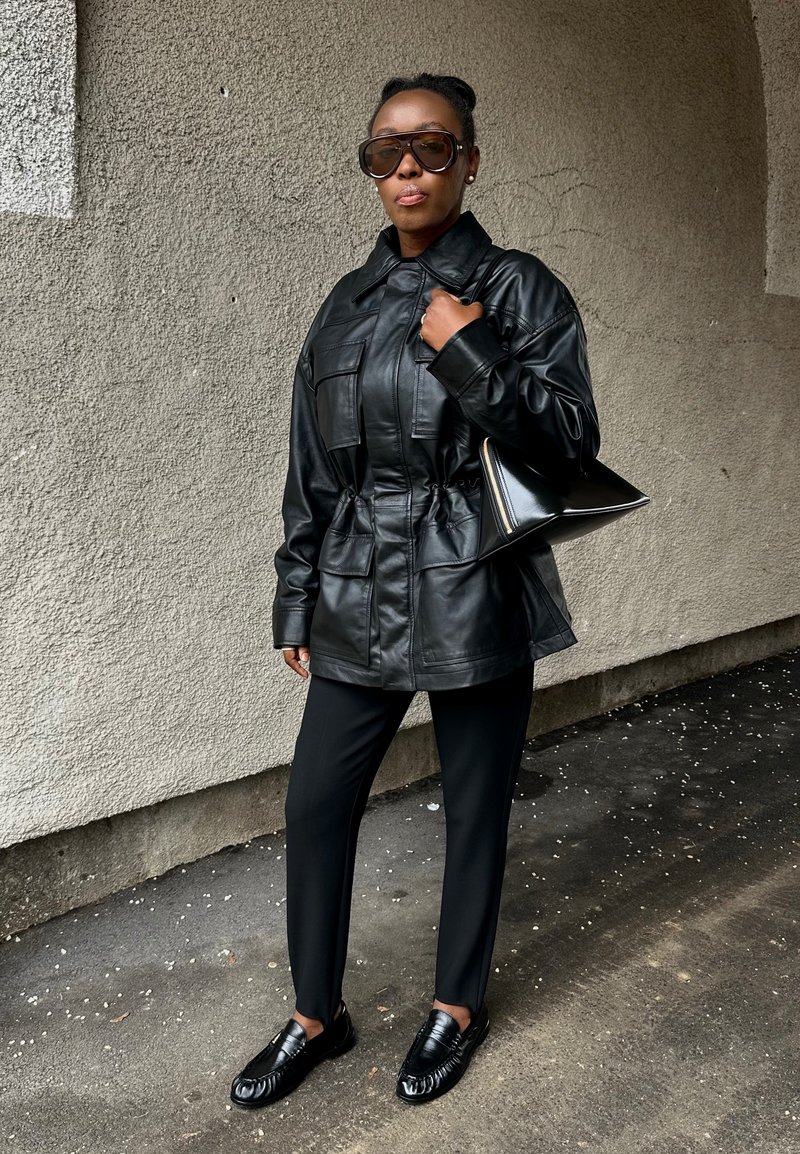 Femme portant une veste et un pantalon en cuir noir, tenant un sac à main noir, debout contre un mur texturé sous une voûte, portant de grandes lunettes de soleil.