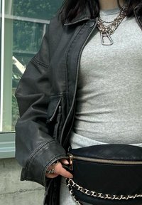 Giacca di pelle nera sopra un top grigio a costine, con una cintura nera trapuntata dotata di una zip dorata e una tracolla a catena. Catene adornano il collo.