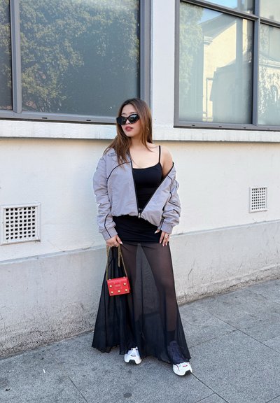 Chaqueta bomber gris claro, vestido negro de tirantes finos, falda negra translúcida, bolso rojo con detalles dorados, zapatillas blancas. Entorno urbano interior.