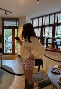 Una persona vestida con una camiseta blanca de gran tamaño y pantalones cortos a rayas rosas se encuentra de pie, sosteniendo una taza, en una cafetería con suelos de madera y grandes ventanales.