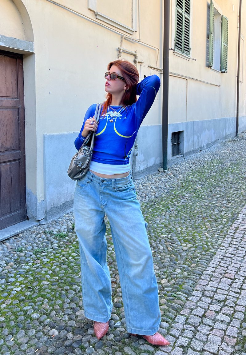 Φωτεινό μπλε μπλουζάκι με μακριά μανίκια και λευκές λεπτομέρειες, oversized ανοιχτό μπλε τζιν παντελόνι και ροζ τακούνια. Κρατάει μια μικρή ασημένια τσάντα.