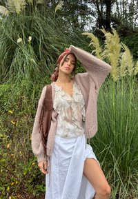 Cárdigan de punto en rosa claro con mangas largas, combinado con una blusa blanca transparente con volantes, y una falda envolvente blanca. Bolso marrón con flecos.