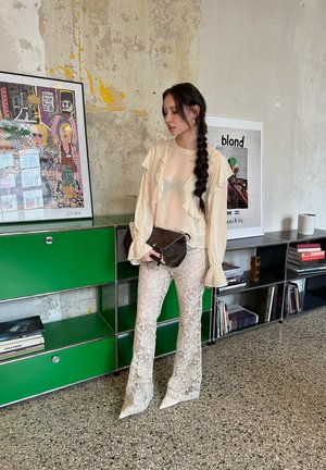 Blusa beige con volantes y mangas largas, combinada con pantalones acampanados de encaje transparente en tonos claros. Bolso de mano marrón oscuro y tacones blancos de punta.