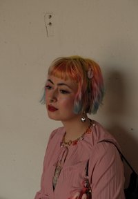 Una persona con cabello en tonos pastel de rosa, azul y amarillo, vistiendo una blusa plisada rosa, un collar colorido y pendientes en forma de corazón.