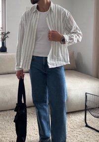 Άνδρας που φοράει λευκό πουκάμισο με ρίγες πάνω από λευκό φανελάκι και μπλε τζιν, κρατώντας μια μαύρη τσάντα, στέκεται σε ένα σαλόνι δίπλα σε ένα μπεζ καναπέ.