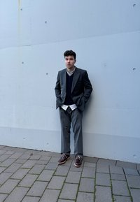 Γκρι σακάκι διπλής όψης, ναυτικό πλεκτό γιλέκο, λευκό πουκάμισο και καφέ παπούτσια στέκονται μπροστά σε ένα ανοιχτό γκρι τοίχο πάνω σε σχεδιασμένο πεζοδρόμιο.