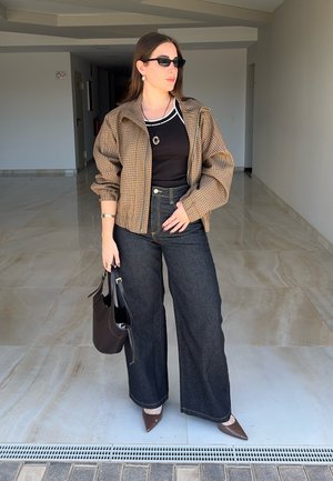 Mujer con gafas de sol, chaqueta marrón a cuadros, top negro, vaqueros de pierna ancha y zapatos marrones puntiagudos sosteniendo un bolso negro en interiores.