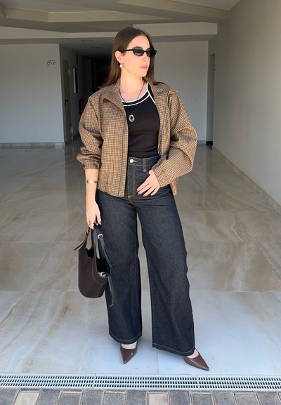 Mujer con gafas de sol, chaqueta marrón a cuadros, top negro, vaqueros de pierna ancha y zapatos marrones puntiagudos sosteniendo un bolso negro en interiores.