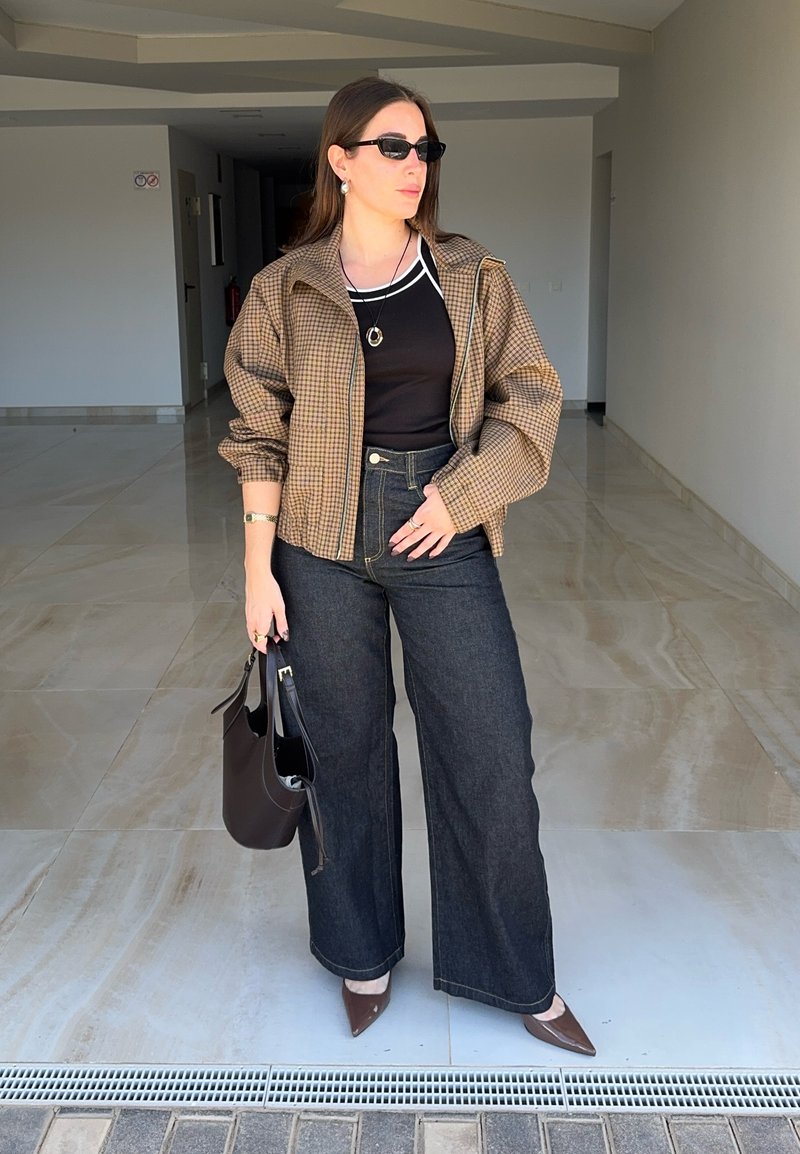 Femme portant des lunettes de soleil, veste marron à carreaux, haut noir, jean à jambes larges et chaussures marron à bouts pointus, tenant un sac à main noir à l'intérieur.