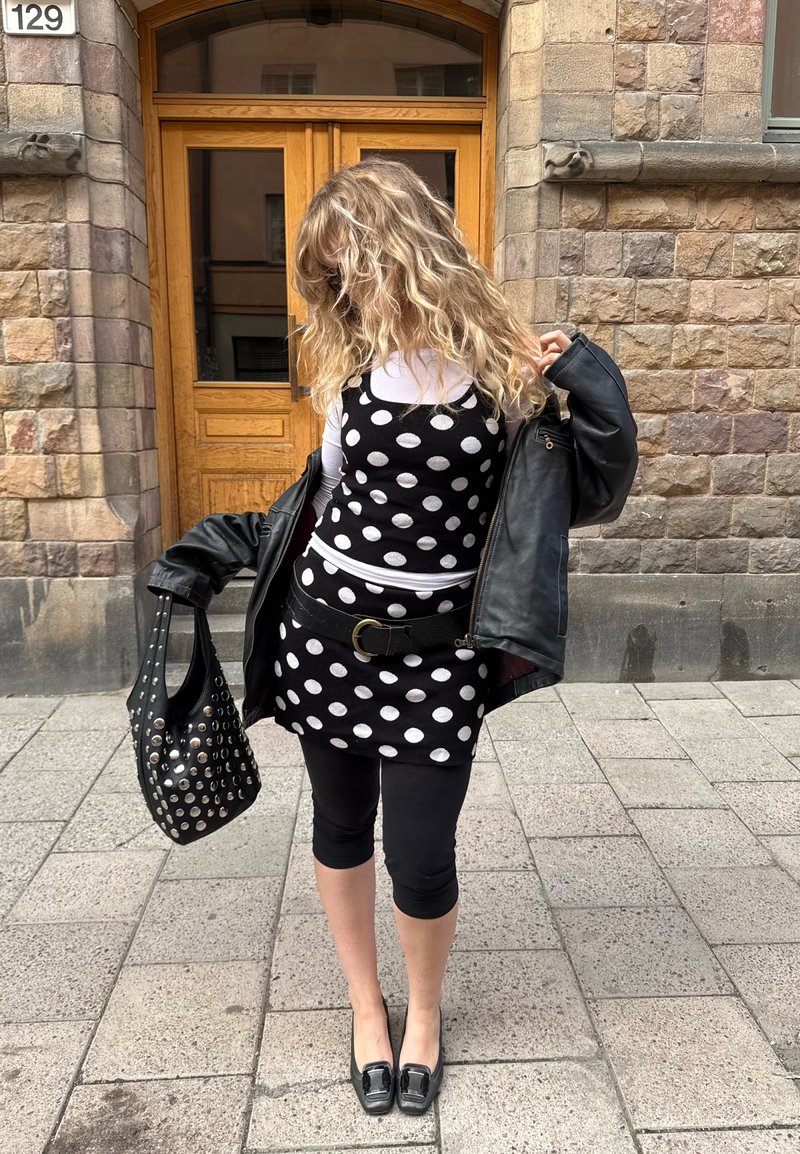 Vestido preto e branco com bolinhas, sobre uma blusa de manga longa branca, combinado com leggings pretas e uma jaqueta de couro. Os acessórios incluem uma bolsa preta com tachas.