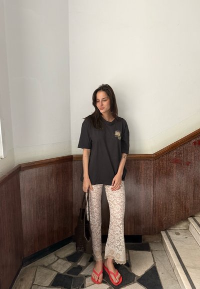 Mujer con cabello oscuro está de pie en una esquina usando una camiseta negra, pantalones blancos con encaje floral, chanclas rojas y sosteniendo una bolsa oscura junto a una escalera.