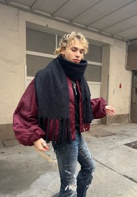 Maroon bomber jacket, black knit scarf with fringes, distressed blue jeans, silver rings, and curly hair. Background shows a concrete wall.