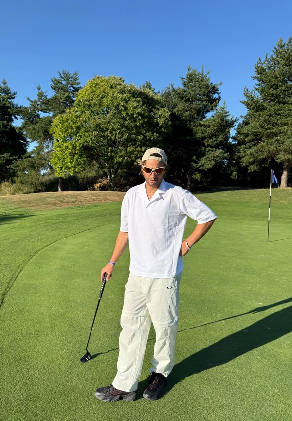 Mand i hvid skjorte og bukser holder en golfputter på en grøn golfbane med træer og et flag i baggrunden.