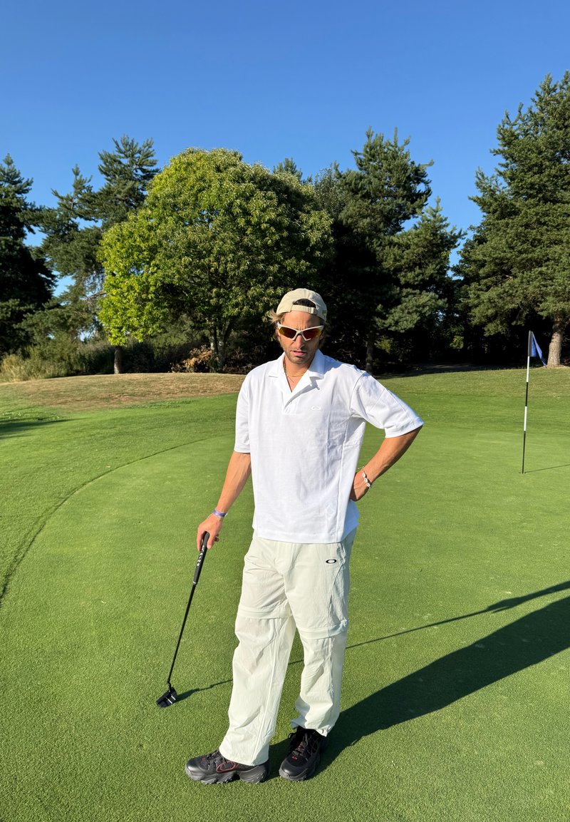 Man in wit hemd en broek houdt een golfputter vast op een groene golfbaan met bomen en een vlaggenstok op de achtergrond.