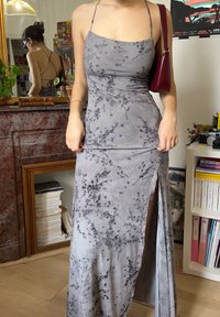 Femme portant une robe nuisette grise à motifs floraux avec des fines bretelles et une fente haute sur la cuisse, tenant le tissu, sac à main rouge sur l'épaule, en intérieur.