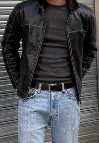 Homme portant une veste en cuir noire par-dessus une chemise côtelée noire, un jean bleu clair et une ceinture noire, posé devant un rideau métallique.