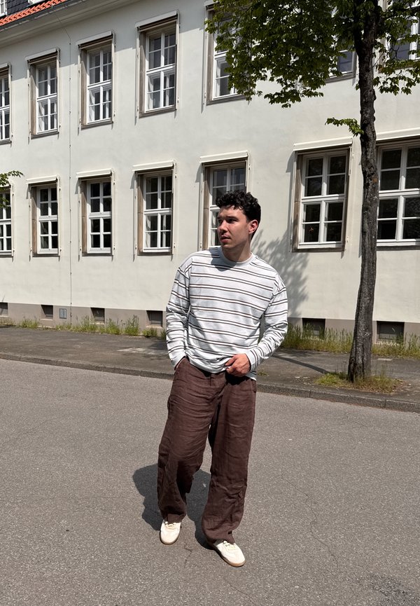 Jeune homme portant une chemise blanche à rayures et un pantalon marron, debout dans une rue devant un bâtiment blanc avec plusieurs fenêtres et un arbre à proximité.