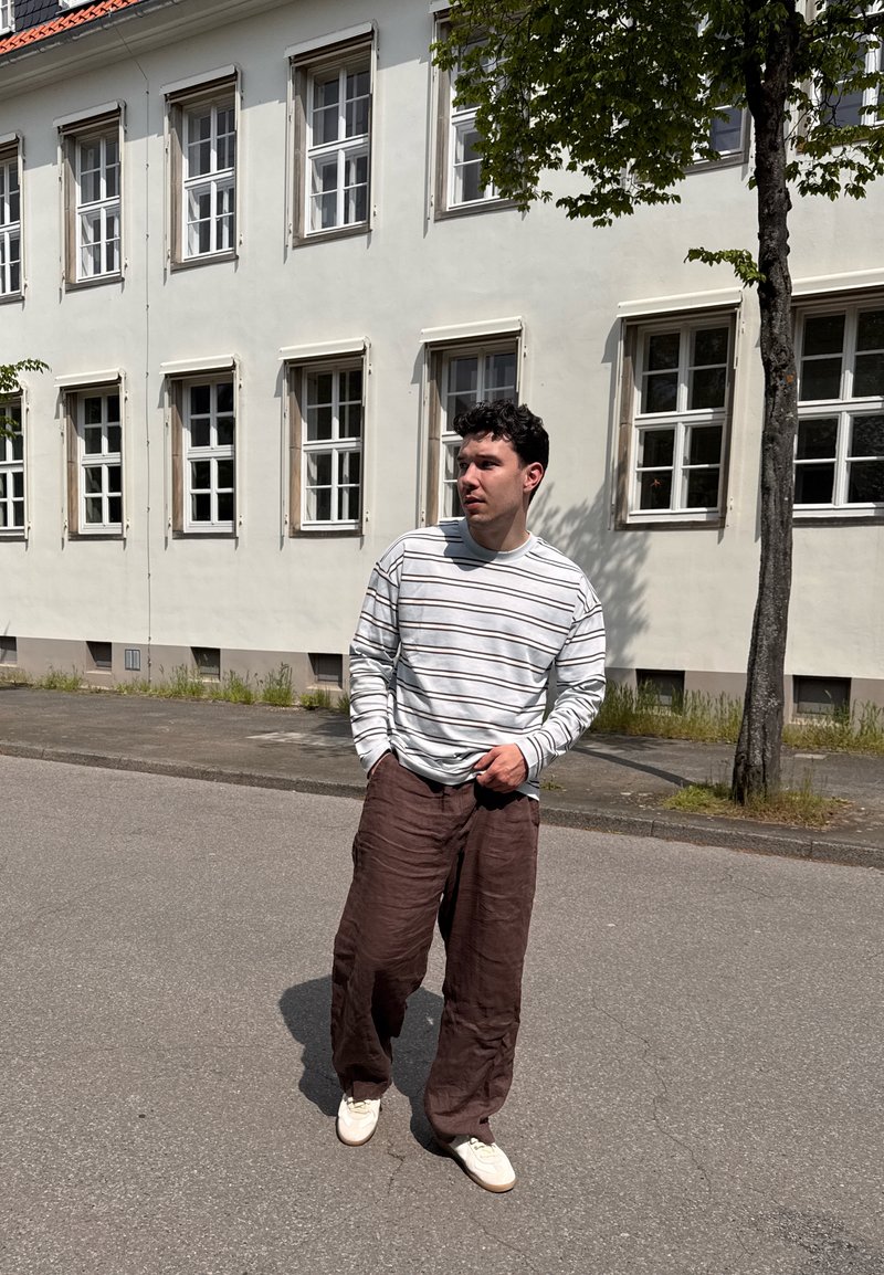 Jeune homme portant une chemise blanche rayée et un pantalon marron, debout dans une rue devant un bâtiment blanc avec plusieurs fenêtres et un arbre à proximité.