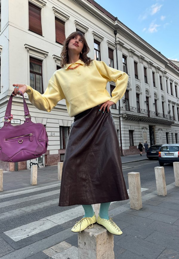 Mujer con suéter amarillo y falda larga marrón está de pie sobre un bloque de hormigón, sosteniendo un bolso morado en una calle de la ciudad con edificios históricos.