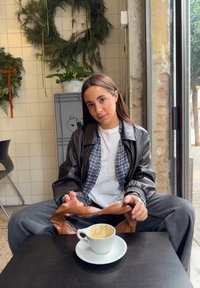 Jeune femme en veste en cuir noire assise à une table de café avec une tasse de café vide, tenant un sac marron, à l'intérieur près d'une fenêtre et de verdure sur le mur.