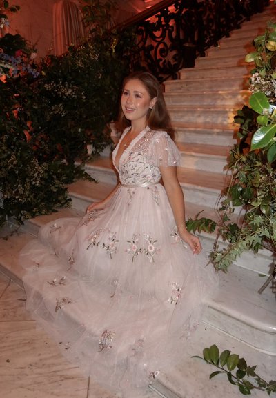 Joven mujer con un vestido blanco bordado con flores sentada en unas escaleras de mármol, rodeada de follaje verde y una barandilla negra ornamentada.