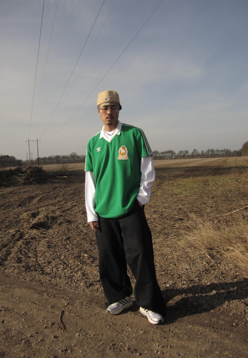Man wearing beige cap, green short-sleeve shirt over white long-sleeve, black pants, and sneakers stands on dirt path by open field under cloudy sky.