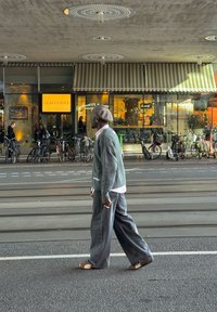 Sivi pleteni kardigan, svijetloružičasta košulja i široke sive traperice. Hoda po pločniku s biciklima u pozadini i kafićem u vidokrugu.