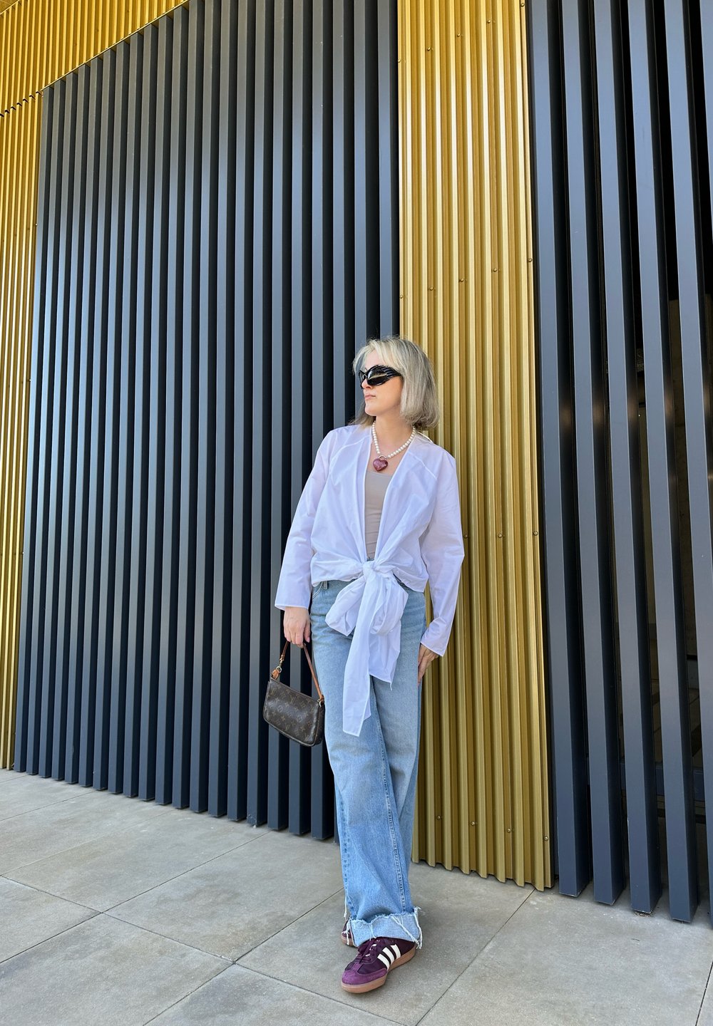 Chemise blanche à nouer à l'avant, haut marron clair, jean bleu avec ourlet effiloché, baskets violettes et blanches, tenant un petit sac à main marron à motif.