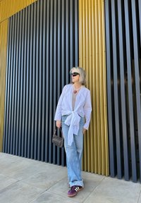 Chemise blanche à nouer à l'avant, haut marron clair, jean bleu avec ourlet effiloché, baskets violettes et blanches, tenant un petit sac à main marron à motif.