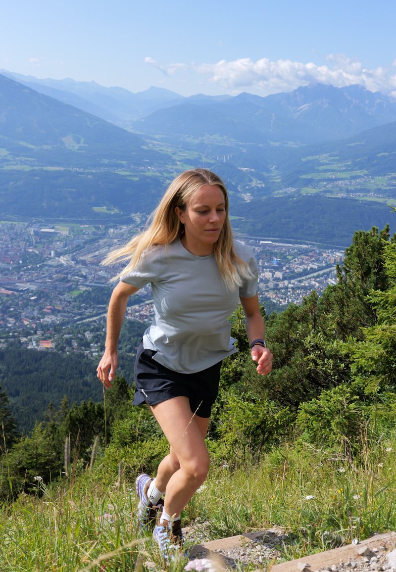 Femme en tenue de sport courant en montée sur un sentier de montagne avec une ville et une chaîne de montagnes visibles en arrière-plan.