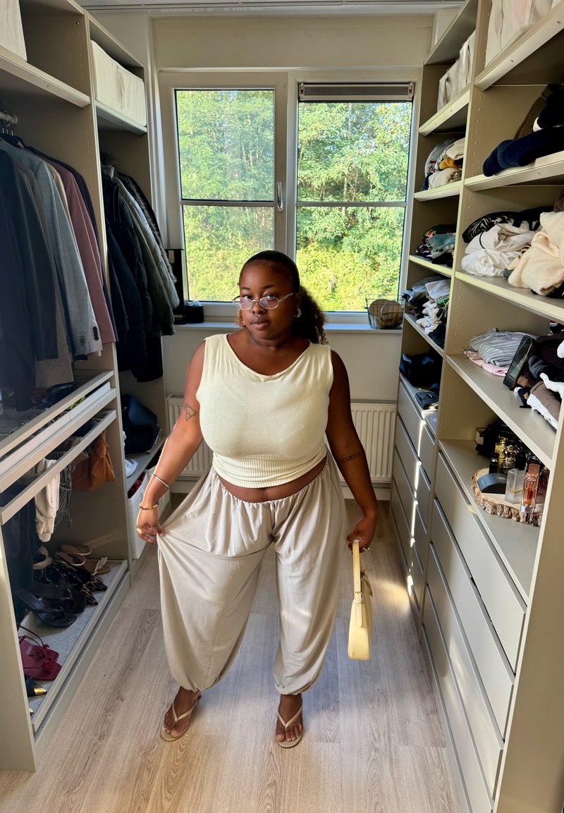 Vrouw draagt beige losse broek en mouwloos topje, houdt een kleine handtas vast en staat in een inloopkast met kleding op planken en hangend.