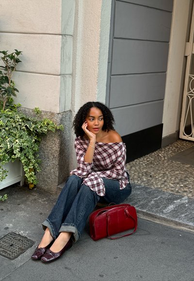 Una camisa de cuadros fuera del hombro, jeans oscuros acampanados, bailarinas burdeos y un bolso a juego, sentada en un suelo de piedra junto a una planta verde.