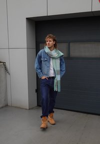 Veste en denim, chemise blanche, jean bleu et bottes couleur beige; enveloppé dans une écharpe vert clair à franges. Debout devant une porte de garage grise.