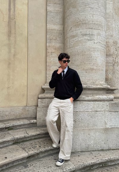 Suéter negro, camisa blanca y pantalones de pierna ancha beige con bolsillos. Zapatillas negras y gafas de sol. Fondo de piedra con columnas texturizadas.