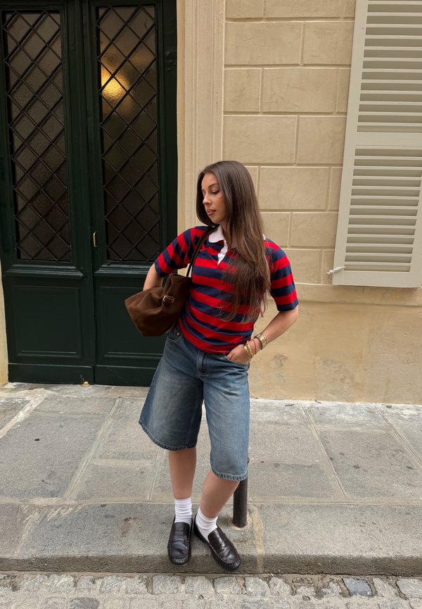 Junge Frau mit langen Haaren in einem rot-navy gestreiften Shirt und Jeansshorts steht auf dem Gehsteig an einer beigen Wand und einer grünen Tür.