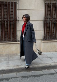 Femme portant un long manteau sombre, une écharpe rouge, des lunettes de soleil, un jean et des mocassins, marchant sur le trottoir en tenant un sac à main à motif par sa chaîne.